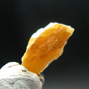 May include: Close-up of a rough, amber-colored gemstone with a textured surface. The stone is mounted on a light-colored, textured base, set against a dark background. The gemstone appears to be a mineral specimen.