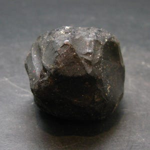 May include: A dark, irregularly shaped stone with a rough, textured surface. The stone is a deep, charcoal color, with subtle variations in tone. The overall shape is rounded, with visible facets and edges. The stone appears to be a natural mineral specimen.