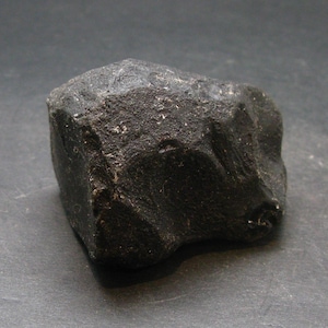 May include: A dark, irregularly shaped rock with a rough, textured surface. The rock is a deep black color and appears to be a natural stone. The image is a close-up shot of the rock against a dark background.