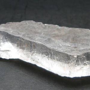 Puede incluir: Una formación cristalina rectangular translúcida con apariencia en capas. El cristal es principalmente transparente con sutiles variaciones de tono, y descansa sobre una superficie oscura. Los bordes muestran una textura rugosa y natural.