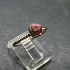 Könnte beinhalten: Ein Ring mit einem kleinen, rohen, dunkelrosa Edelstein, der in eine polierte, silberfarbene Metallschiene gefasst ist. Der Stein hat eine unregelmäßige, texturierte Oberfläche und wird von einer einfachen Zargenfassung gehalten.