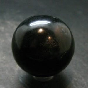 Peut inclure: Une sphère noire solide et lisse avec une surface polie et réfléchissante. L'objet est parfaitement rond et présente une finition brillante qui capte la lumière, créant un reflet blanc vif sur sa partie supérieure sur le fond sombre.