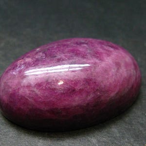 Op de afbeelding: Een ovaalvormige cabochon edelsteen met een gepolijst, glad oppervlak. De steen heeft een diep magenta en paars kleurenpalet met wervelende, gevlekte patronen en natuurlijke insluitingen die zichtbaar zijn door de afgeronde koepelvorm.