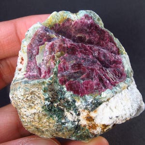 May include: A rough, irregularly shaped rock with a large, pink-red crystal embedded in it. The crystal is surrounded by a matrix of green and white minerals.