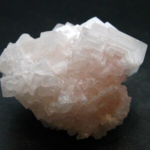 May include: A close-up of a pale pink halite crystal cluster. The crystal structure is cubic, with visible stepped formations and a translucent appearance. The mineral has a slightly rough texture and is set against a dark background.