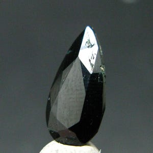 Puede incluir: Una piedra preciosa negra en forma de pera con una superficie facetada. La piedra presenta una parte superior puntiaguda y una base redondeada, con múltiples superficies planas y pulidas que reflejan la luz. El color negro es uniforme.