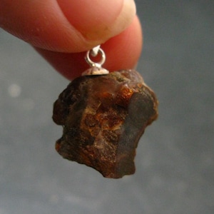 May include: A raw, dark brown amber pendant with a rough, irregular texture and a polished surface. The stone is attached to a small, silver-colored metal bail at the top, allowing it to be suspended from a chain or cord for use as jewelry.