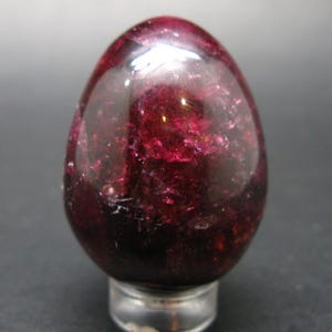 May include: A polished, egg-shaped gemstone in deep red hues. The stone has a glossy surface with internal crystalline structures, mounted on a small, silver-coloured base. The overall appearance is smooth and reflective, with variations in colour.