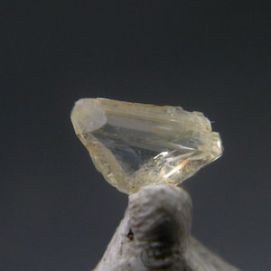 Puede incluir: Un cristal translúcido de color amarillo claro con una forma geométrica facetada. El cristal está montado sobre una pequeña base texturizada de color blanquecino. El fondo es gris oscuro liso.