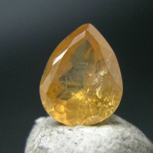 May include: A teardrop-shaped, faceted gemstone with a golden yellow color. The stone has a unique, cloudy appearance with internal inclusions.