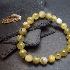May include: A bracelet made of light yellow and green gemstone beads. The beads are round and have a smooth, polished finish. The bracelet is on a dark grey background.