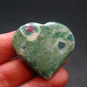 Könnte beinhalten: Ein herzförmiger Edelstein mit grüner Grundfarbe, gesprenkelt mit dunkleren grünen und weißen Flecken. Ein kleiner, leuchtend roter Einschluss ist sichtbar. Der Stein wird in einer Hand gehalten.