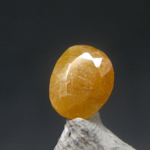 May include: A faceted, oval-shaped, golden-yellow gemstone. The gem is mounted on a small, rough, gray stone, set against a dark gray background. The gem's facets reflect light, creating bright highlights.
