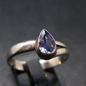 May include: A silver ring featuring a pear-shaped, deep blue gemstone. The ring's band has a split design, with a smooth, polished finish. The gemstone is set in a bezel setting, enhancing its color and shape.