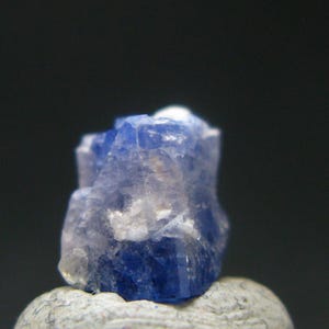 May include: A close-up of a blue and white crystal formation. The crystal has a rough, textured surface and is set against a dark background. The crystal's color transitions from a deep blue to a lighter, almost white hue.