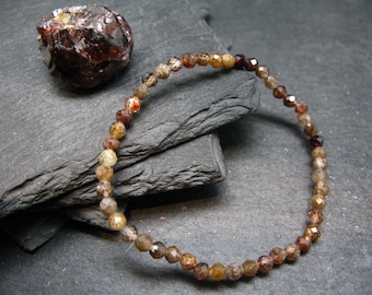 May include: A beaded bracelet with small, faceted, brown and amber-colored beads. A rough, dark brown gemstone sits nearby. The bracelet is strung on a clear, elastic cord. The beads have a sparkling, reflective quality.