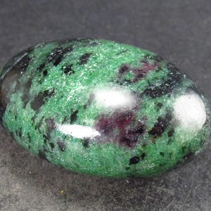 Peut inclure: Une pierre précieuse ovale de couleur verte et noire avec une surface lisse et polie. La pierre a un aspect marbré avec des inclusions vert foncé, noires et rouges.