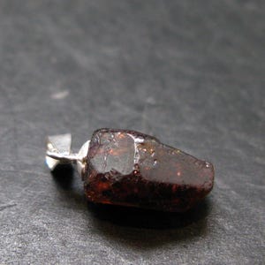 May include: A raw, dark red gemstone pendant with a silver bail. The gemstone has a rough, natural texture and is irregularly shaped. The pendant is set against a dark background.