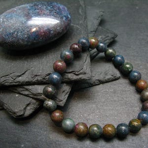 May include: A dark blue, green and brown gemstone bracelet with round beads. The bracelet is lying on a stack of slate tiles.