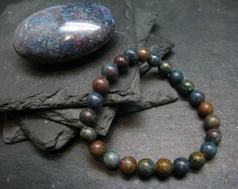 May include: A dark blue, green, and brown gemstone bracelet with round beads. The bracelet is lying on a stack of slate tiles.