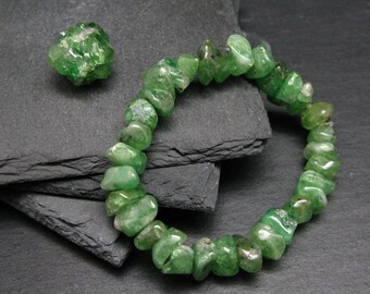 May include: A green gemstone bracelet made of small, irregular shaped beads. The bracelet is on a dark gray surface. There is a single green gemstone next to the bracelet.