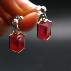 May include: A pair of silver-toned earrings with a geometric design. Each earring features a rectangular, faceted, deep red gemstone set in a gold-colored frame. The earrings are suspended from a small, decorative silver stud.