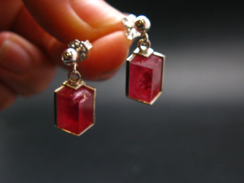 May include: A pair of silver-toned earrings with a geometric design. Each earring features a rectangular, faceted, deep red gemstone set in a gold-colored frame. The earrings are suspended from a small, decorative silver stud.