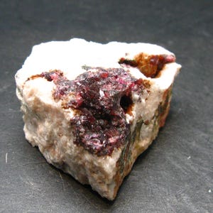 Puede incluir: Un espécimen mineral en bruto con una mezcla de estructuras cristalinas blancas, marrón rojizo y rojo oscuro. La roca tiene una superficie texturizada con variaciones visibles en color y composición, mostrando formaciones geológicas naturales.