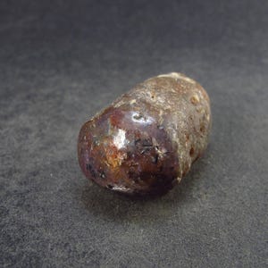 May include: A dark brown, irregularly shaped, polished stone with a smooth, shiny surface. The stone has a reddish-brown hue and appears to be translucent.