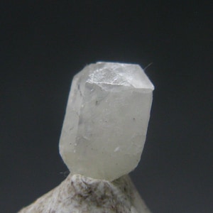 Könnte beinhalten: Ein durchscheinender, hellfarbener Kristall mit facettierter Oberseite und rechteckiger Form. Der Kristall liegt auf einer rauen, hellfarbigen Oberfläche vor einem dunkelgrauen Hintergrund.