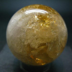 Peut inclure: Une sphère de cristal translucide jaune doré. La surface polie reflète la lumière, révélant des textures internes et des variations de couleur. La sphère est un objet décoratif, peut-être pour l'exposition ou la méditation.