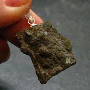May include: A raw, irregularly shaped pendant featuring a rough, textured surface with shades of brown and green. The pendant is suspended from a small silver bail. The natural stone has small, sparkling inclusions.