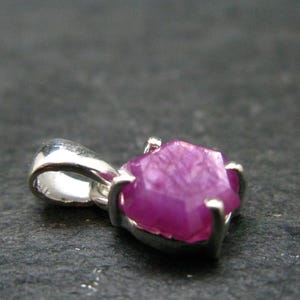 May include: A pendant featuring a raw, hexagonal-shaped pink ruby gemstone set in a silver-toned metal prong setting. The gemstone displays a vibrant magenta hue with visible internal crystalline structures and a polished, reflective bail.