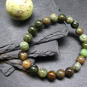 May include: A beaded bracelet featuring round, polished beads in shades of green, brown, and tan. The bracelet is displayed on a dark gray, textured surface, with a matching sphere in the background. The bracelet is a piece of jewelry.