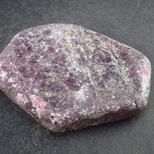 Pode incluir: Uma gema rosa e cinza de forma irregular e bruta. A pedra tem uma superfície áspera e texturizada e é opaca.