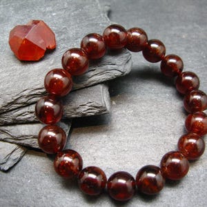 Puede incluir: Un brazalete de cuentas con cuentas grandes, pulidas, redondas, de color rojo oscuro. Una gema facetada, de color naranja rojizo, se encuentra cerca. La joyería se exhibe sobre una superficie oscura y texturizada, mostrando el rico color y brillo de las gemas.