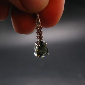 May include: A gold-colored pendant with a pear-shaped, dark green gemstone. The pendant is held between fingers against a dark gray background. Small, reddish-brown stones are set above the main gem.