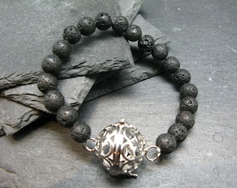 Puede incluir: Un brazalete de cuentas de roca volcánica negra con un dije de jaula de plata. El brazalete está sobre un fondo de pizarra gris.