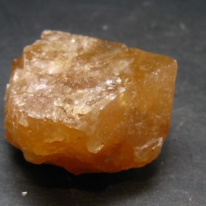 May include: A rough, uncut, amber-colored gemstone with a crystalline structure. The stone has a slightly translucent appearance, with variations in color and texture. The surface reflects light, creating highlights and shadows.