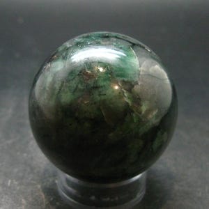 May include: A polished spherical stone featuring a deep forest green colour with dark inclusions and translucent patches. The surface of the sphere is smooth and reflective, showing natural mineral patterns throughout the rounded form.