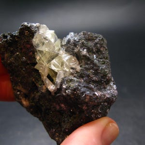 Puede incluir: Una formación rocosa oscura y rugosa con un grupo de cristales translúcidos de color amarillo claro. Los cristales tienen forma geométrica y están agrupados, contrastando con la matriz oscura y texturizada. La roca se sostiene en una mano.