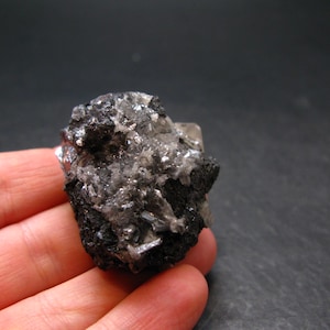 May include: A close-up of a mineral specimen featuring a cluster of dark, crystalline structures with reflective, silvery-gray accents. The mineral has a rough, textured surface and is held in a hand against a dark background.