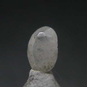 May include: A clear, faceted gemstone with a rounded shape. The gem is set on a small, light-colored base, and the background is a dark gray. The gemstone has a slightly cloudy appearance with subtle inclusions.