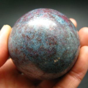 May include: A smooth, round, blue and red gemstone sphere. The sphere has a speckled appearance with a mix of blue, red, and gray colors.