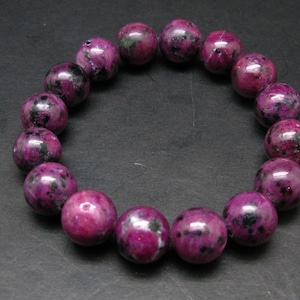 Op de afbeelding: Een armband gemaakt van ronde, gepolijste kralen. De kralen zijn diep magenta van kleur met zwarte en groene spikkels. De armband ligt op een donkergrijze ondergrond.