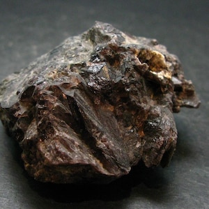 Puede incluir: Un espécimen mineral en bruto de color marrón oscuro con una estructura cristalina irregular. La superficie presenta facetas angulares afiladas y tonos tierra profundos con reflejos brillantes sobre la formación rocosa texturizada.
