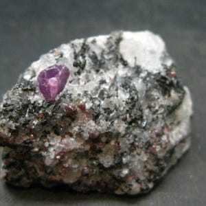 May include: A small, dark gray rock with a bright purple ruby crystal embedded in it. The ruby is faceted and has a sharp point.