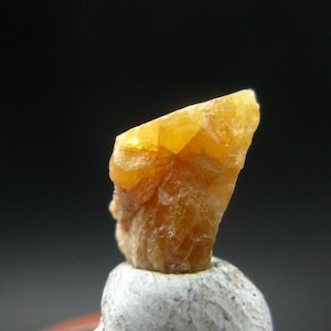 May include: A close-up of a rough, orange-yellow mineral specimen. The crystal has a jagged, irregular shape and sits atop a small, light-colored rock. The background is a dark gray.