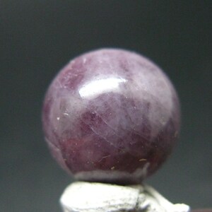 Può includere: Una pietra preziosa sferica lucidata di una tonalità viola e magenta profonda, caratterizzata da venature interne naturali e una superficie liscia e riflettente. La pietra mostra varie sfumature di viola e rosa scuro.