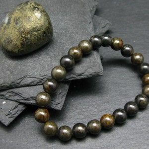 Puede incluir: Un brazalete hecho de cuentas de pirita negra y dorada. El brazalete está sobre una superficie de pizarra negra con una piedra de pirita dorada.